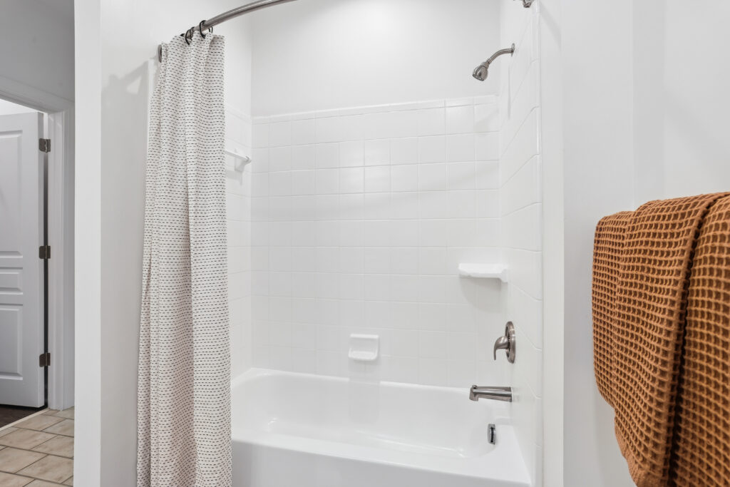 A clean, white bathroom with a bathtub and shower combo. A patterned shower curtain is partially closed, and a brown textured towel hangs on a rack next to the tub. The walls and tiles are white.