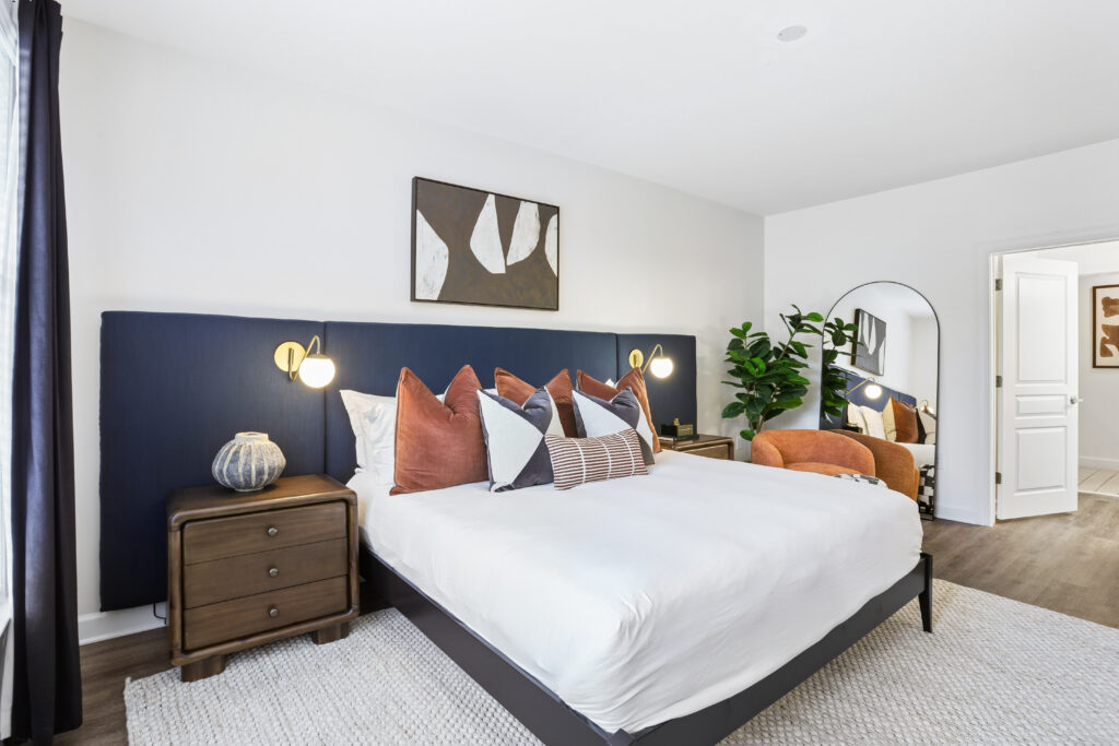 A modern bedroom with a large bed, navy-blue headboard, decorative pillows, wooden nightstands, wall art, a round mirror, an orange chair, a potted plant, and natural light from a window with dark curtains.