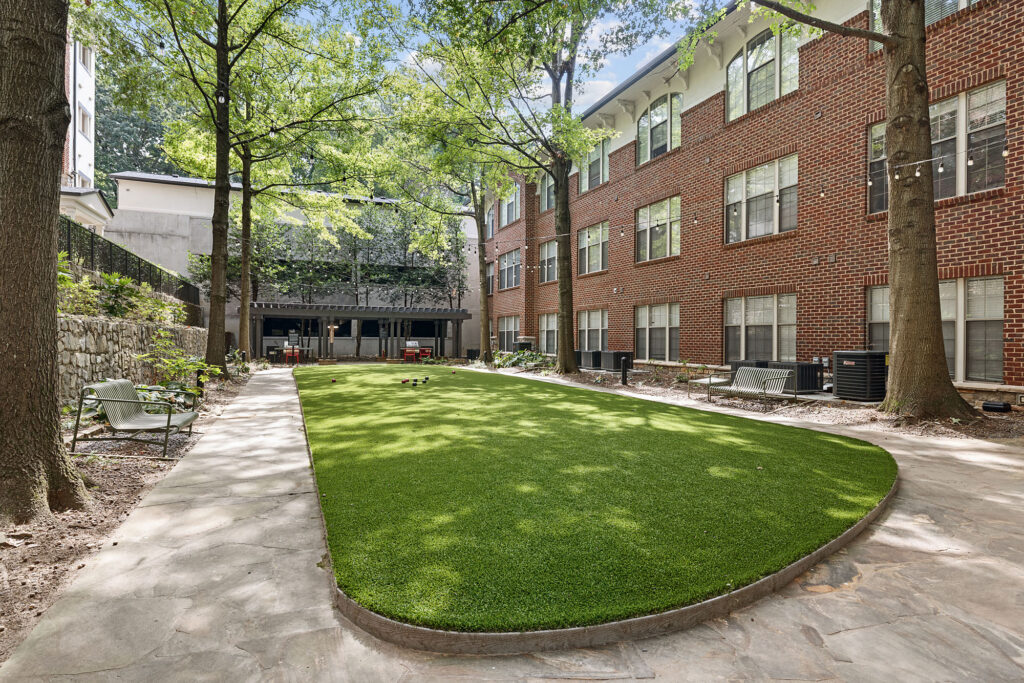 A rectangular artificial turf lawn set between a brick apartment building and a stone retaining wall, with a bocce ball court at the far end and stone pathways surrounding the green.