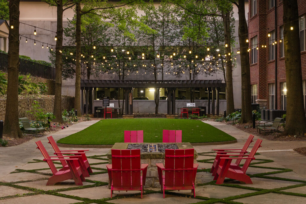 A courtyard with a stone fire pit surrounded by six red Adirondack chairs on a patio with a central patch of artificial turf and string lights hanging above.