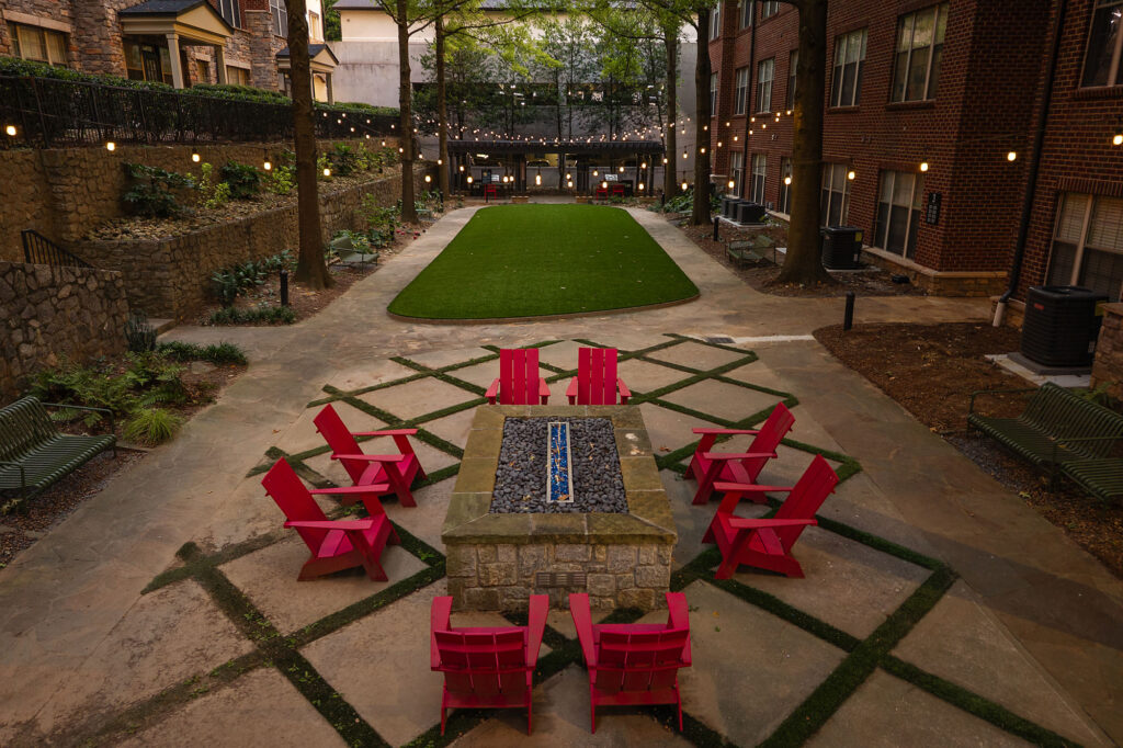 A stone fire pit surrounded by red Adirondack chairs in a landscaped courtyard with a rectangular patch of turf and string lights.
