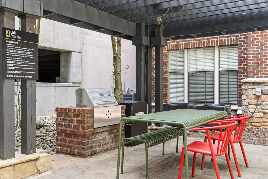 An outdoor patio with a brick grill station, a green metal dining table, and red chairs under a pergola, with a sign detailing grill policies.