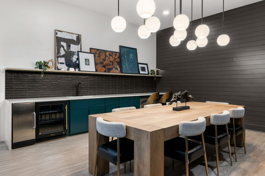 A large wooden dining table with six chairs sits in a room featuring a dark shiplap wall, a built-in kitchen counter with teal cabinets, and a cluster of spherical pendant lights hanging above.