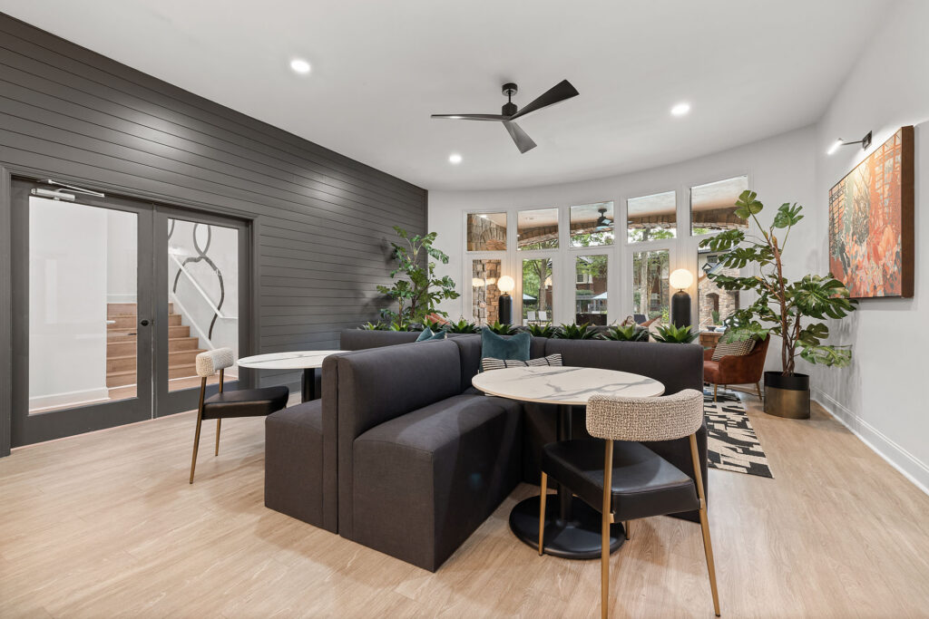 A lounge area featuring dark gray booth seating arranged around round marble tables with modern chairs, set against a dark shiplap wall and large windows.