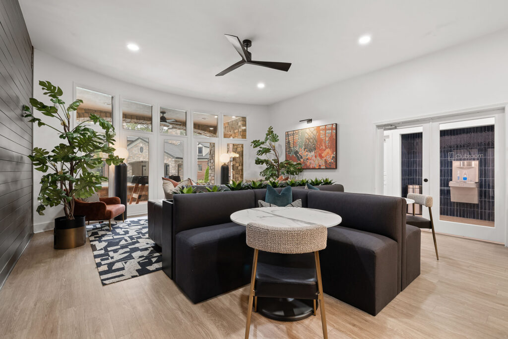 A common area featuring a dark grey sectional sofa, a round marble-topped table, and a hallway view leading to a clubroom with a water dispenser.