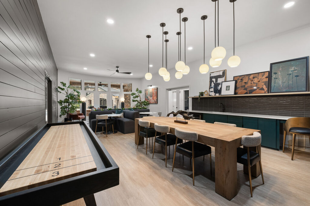 A long wooden dining table with six chairs sits in a room featuring a shuffleboard table, a kitchen counter with dark green cabinetry, and several pendant lights hanging from the ceiling.
