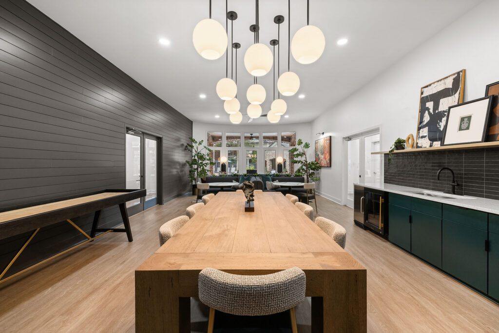 Long wooden dining table with upholstered chairs in a room featuring a dark shiplap wall, a wet bar, and a cluster of spherical pendant lights.