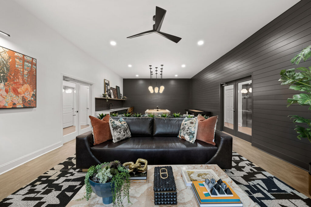 A modern living room with a black leather sofa featuring patterned and solid orange pillows, set before a dining area with a long table and charcoal-colored shiplap walls.