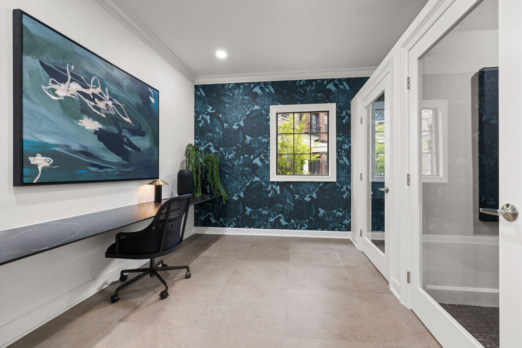 A home office with a dark wall featuring floral wallpaper, a floating desk with a black mesh chair, and a large abstract painting on the wall.