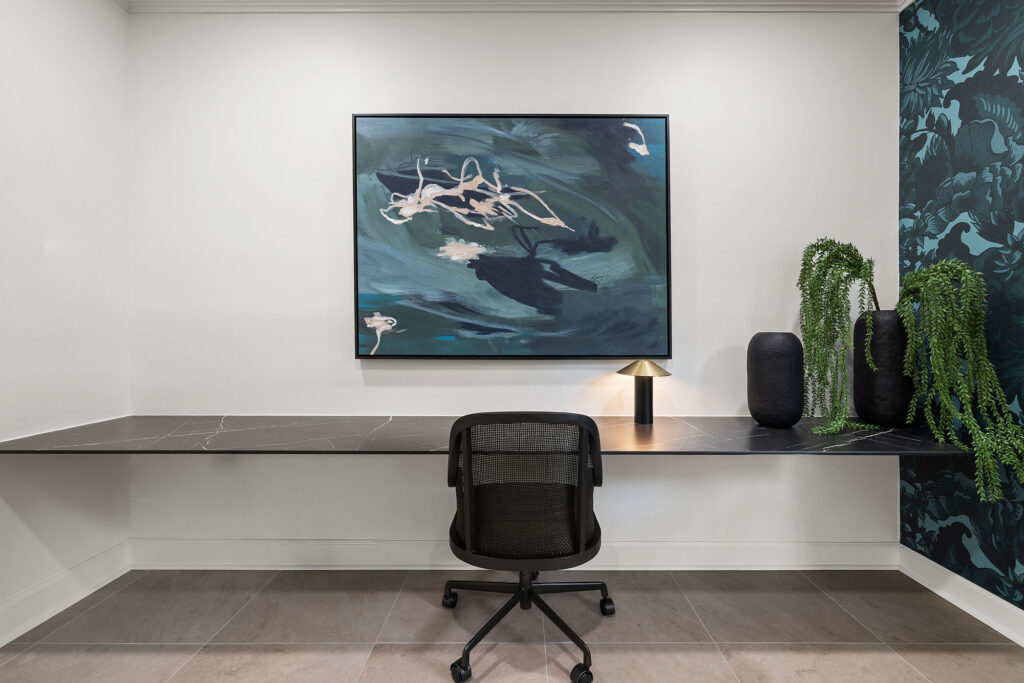 A modern home office featuring a long black desk, a black mesh office chair, and a large abstract painting on the wall.