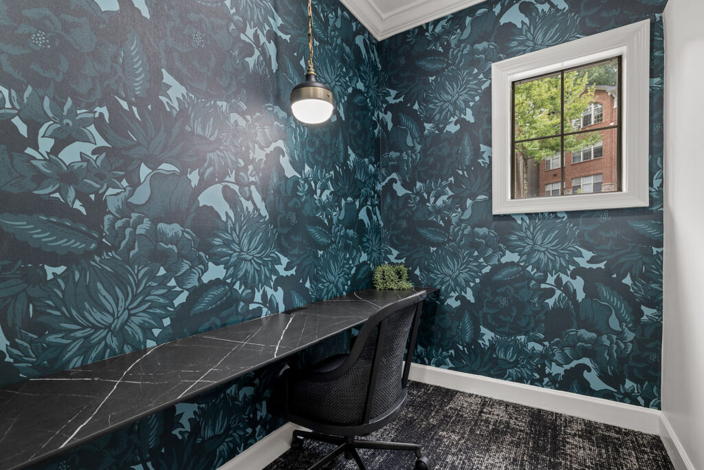 A desk nook with dark blue floral wallpaper, a black marble-look desk, a mesh office chair, and a pendant light.