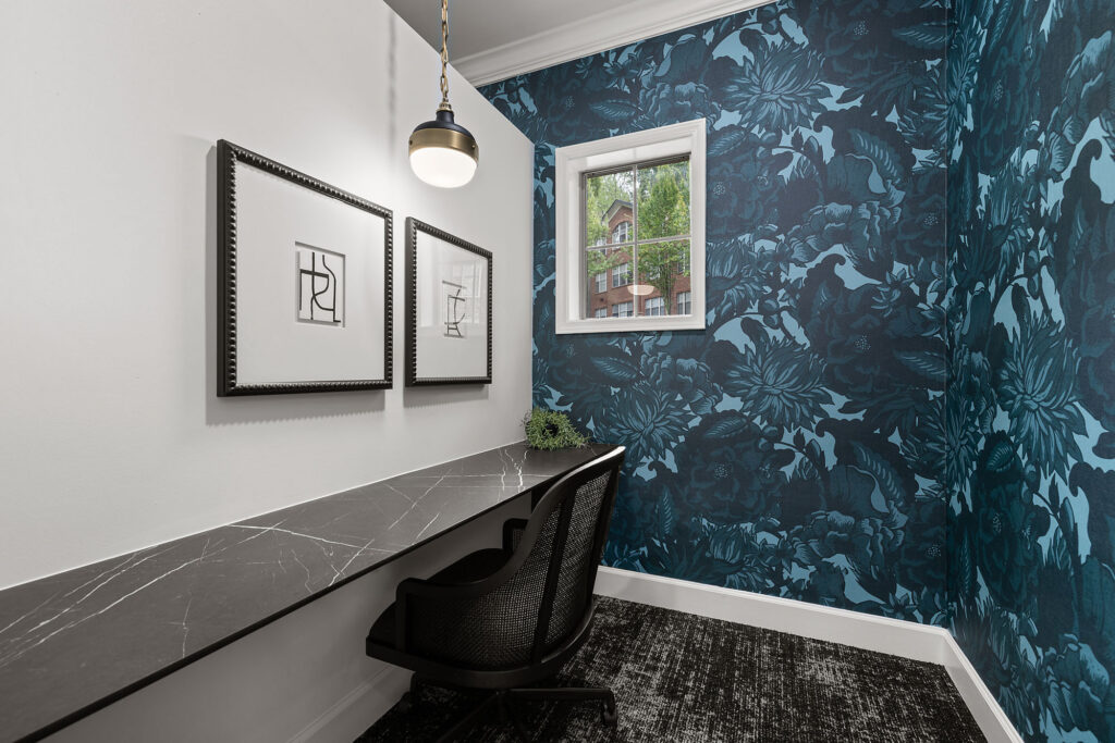 A small home office nook featuring a long marble desk, a black mesh chair, two framed abstract line drawings on a white wall, and dark blue floral wallpaper.