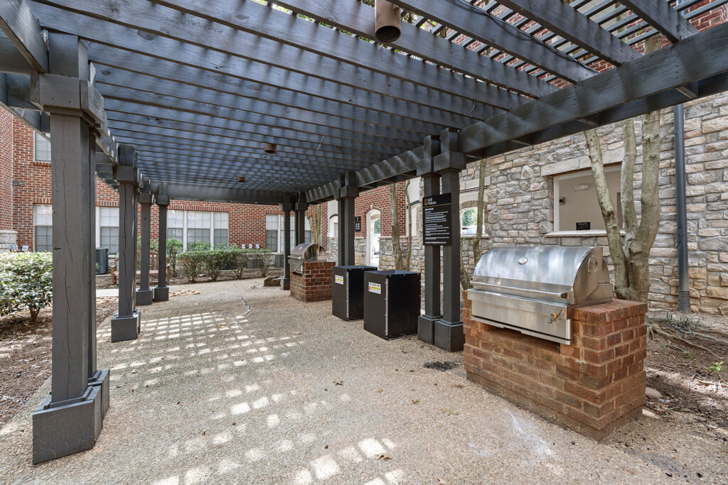 Two stainless steel grills set into brick bases under a long wooden pergola in a courtyard.