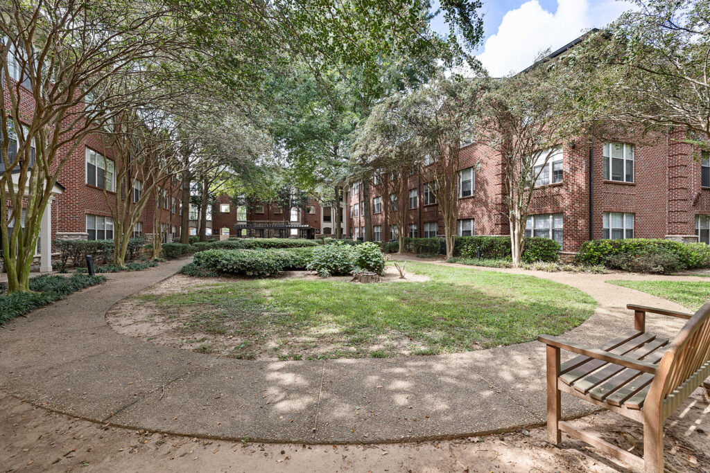 A curved stone pathway surrounds a central grassy courtyard between two red brick apartment buildings, with a wooden bench in the foreground.