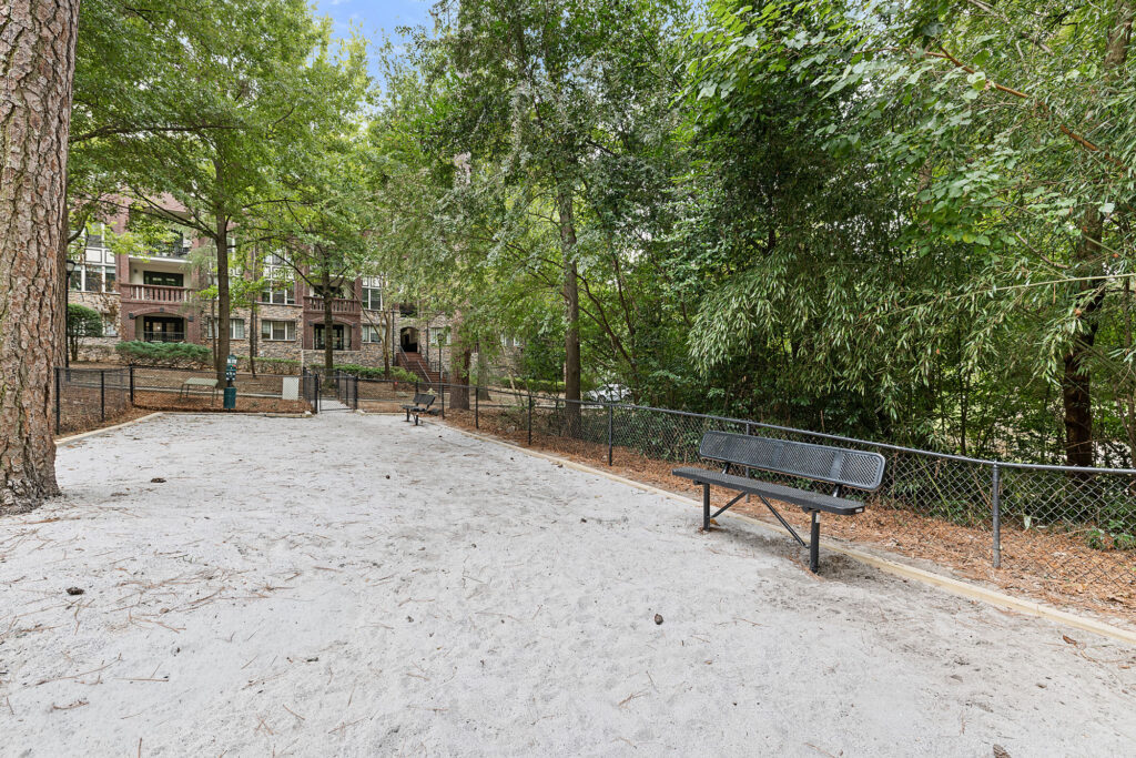 A fenced dog park with a sandy surface, two black metal benches, and a large brick apartment building in the background.