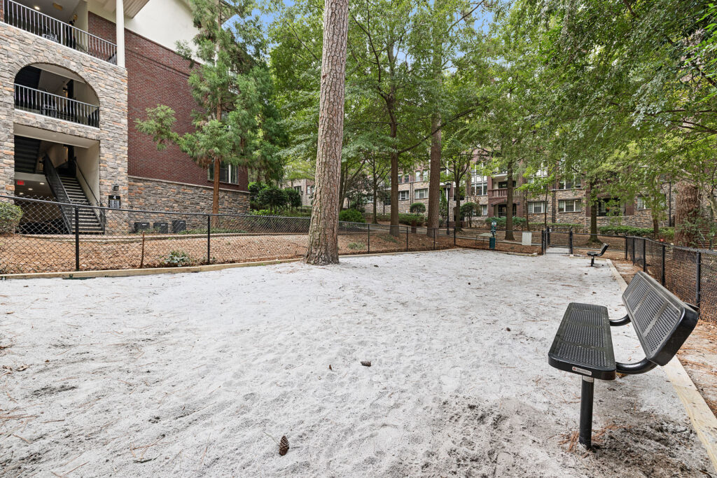A sandy dog park enclosed by a chain-link fence, featuring a metal bench and surrounding trees near a multi-story brick apartment building.