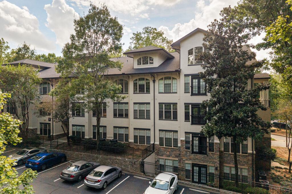 A four-story apartment building with a white stucco and stone facade, surrounded by trees, with parked cars in the foreground.