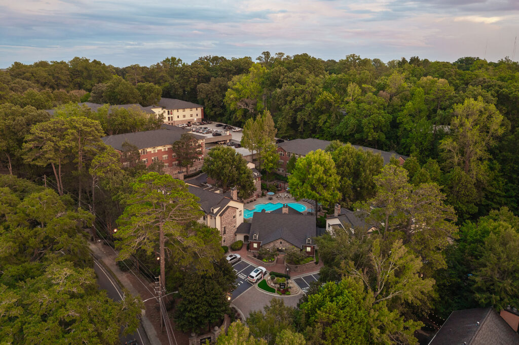 Aerial view of an apartment complex nestled in a forest, featuring several multi-story buildings, a central swimming pool, and parking areas.