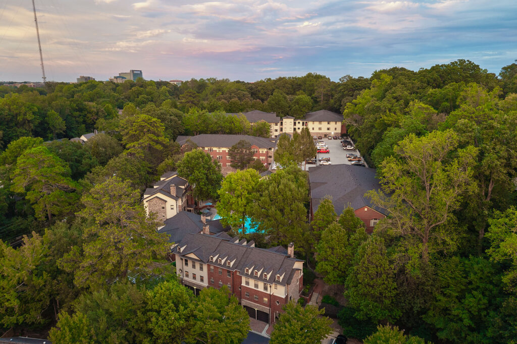Aerial view of an apartment complex nestled in a dense forest, featuring multiple multi-story brick buildings, a parking lot, and a swimming pool.