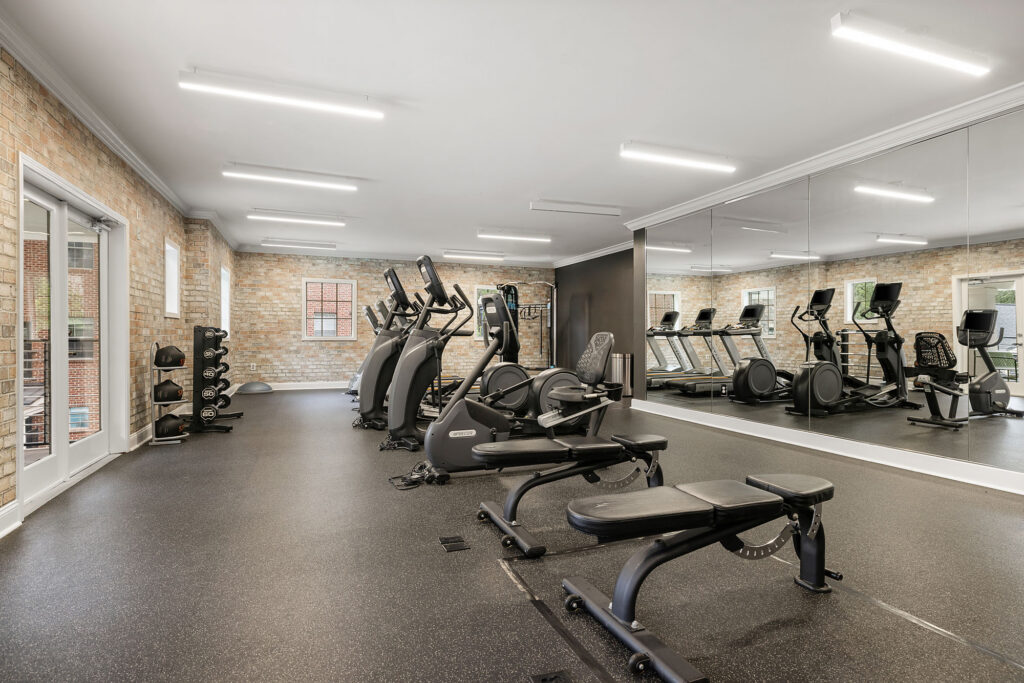 A fitness center with weight benches, elliptical machines, and treadmills, featuring a mirrored wall and brick accents.