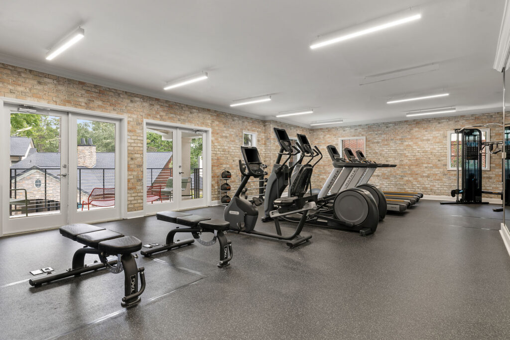 A home gym featuring two weight benches in the foreground, followed by several elliptical machines and treadmills in front of a brick wall with glass doors leading to a balcony.