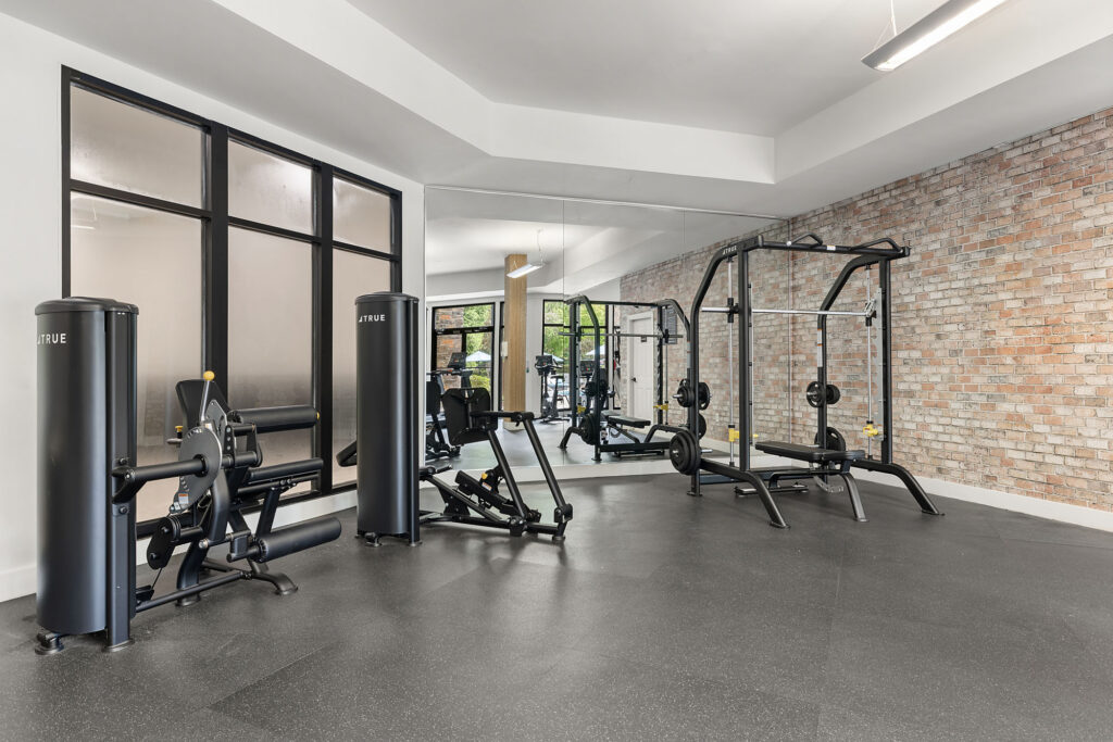 A gym with a leg extension machine and a Smith machine in front of a mirrored wall and brick accent wall.