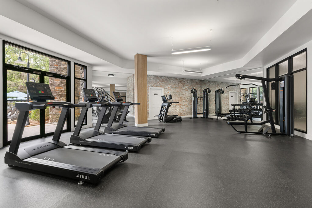 A fitness center featuring several treadmills, a weight machine, and a rack of dumbbells against a brick wall.