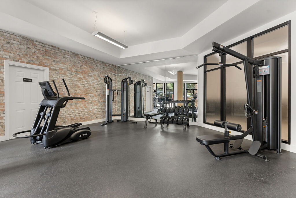 A fitness center featuring a treadmill, a cable machine, a weight bench, and a rack of dumbbells in front of a brick wall and mirrors.