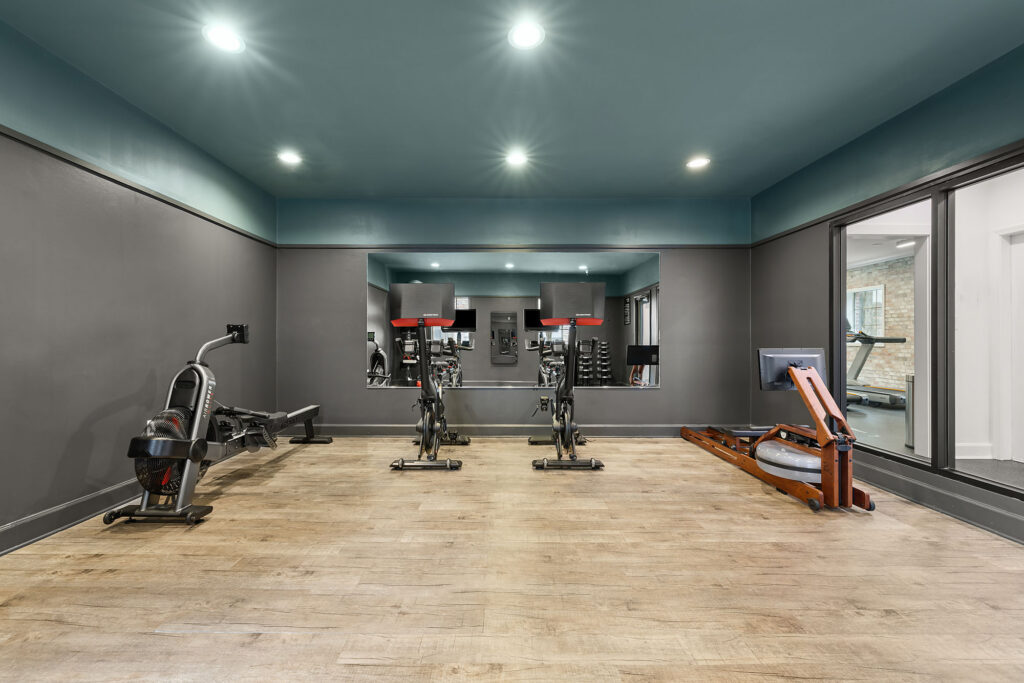 Home gym featuring two stationary bikes and two rowing machines on a wood floor, with a large wall mirror reflecting the equipment.