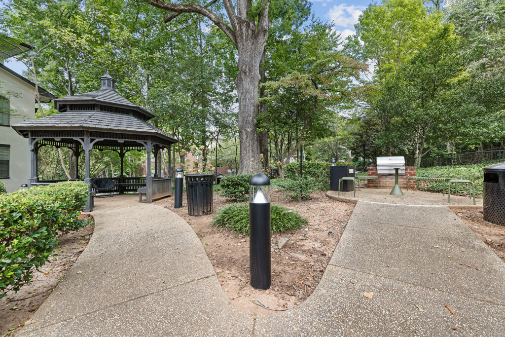A concrete path forks toward a wooden gazebo on the left and a grill station on the right in a wooded area.
