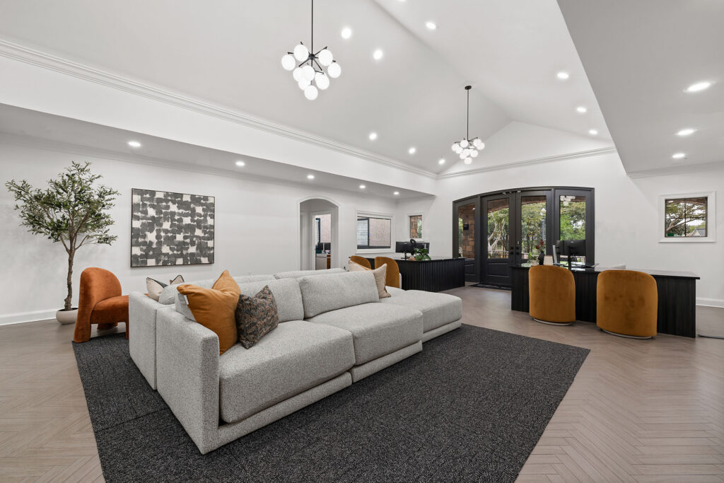 A modern open-concept room features a large gray sectional sofa on a dark rug, with a workspace area containing two desks and mustard-colored chairs in the background.
