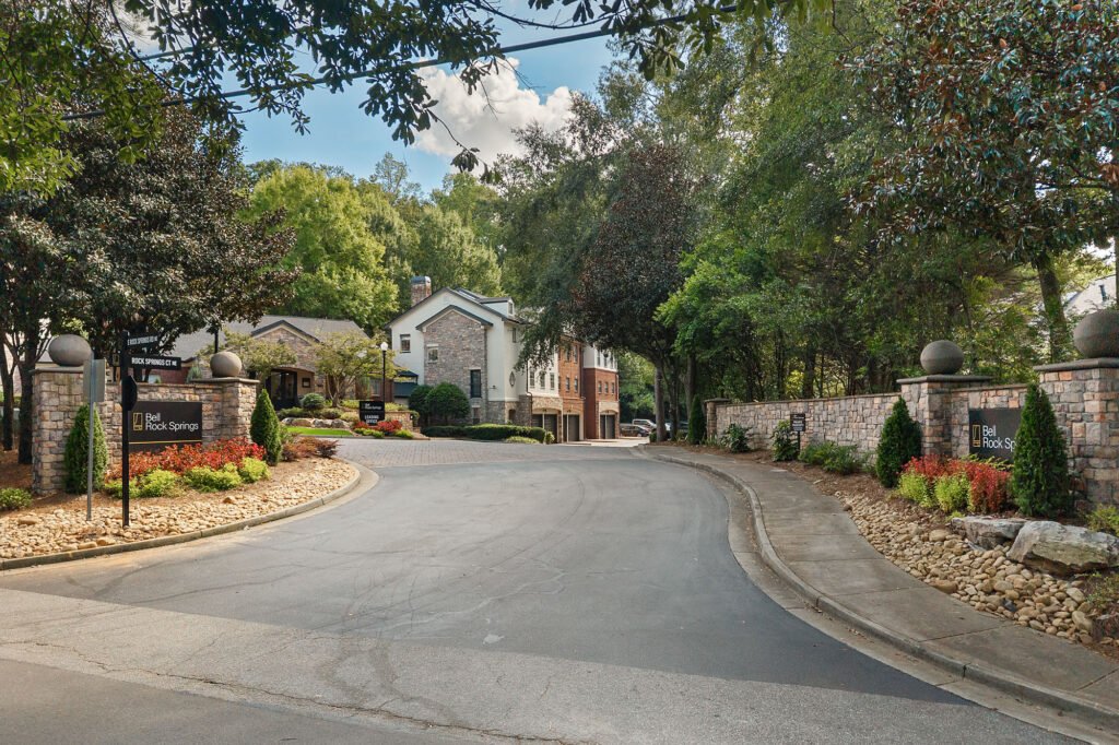 The entrance to Bell Rock Springs apartment community, featuring stone pillars with signage on either side of a paved road leading to residential buildings nestled among trees.