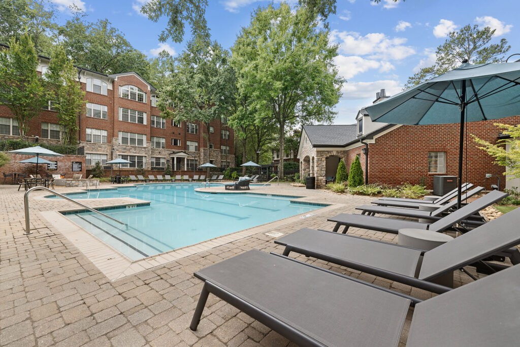 A swimming pool with lounge chairs and umbrellas on a brick patio in front of a multi-story brick apartment building.