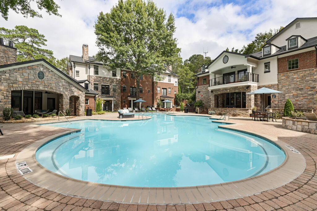 A large, curved swimming pool surrounded by a brick patio and residential buildings with stone and brick exteriors.