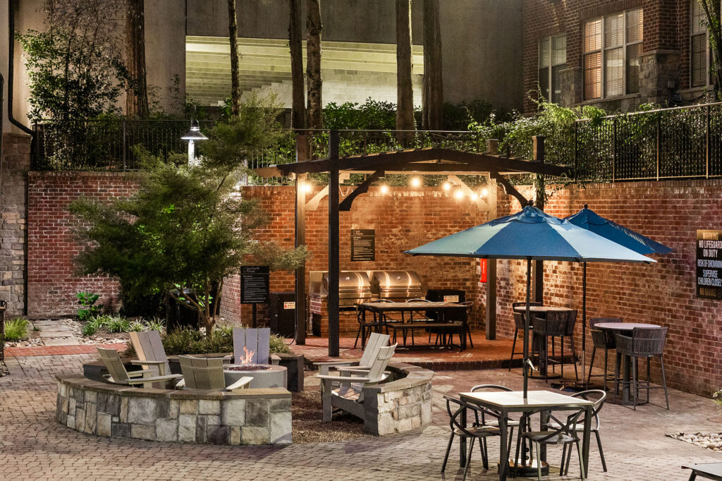 An outdoor courtyard with a fire pit, patio seating, a blue umbrella, and a covered grilling station against a brick wall.