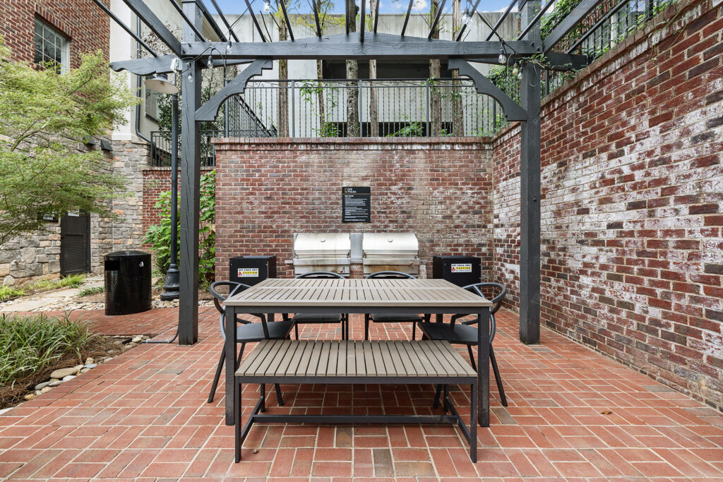Outdoor patio with a metal table, benches, and chairs set in front of two stainless steel grills against a brick wall.