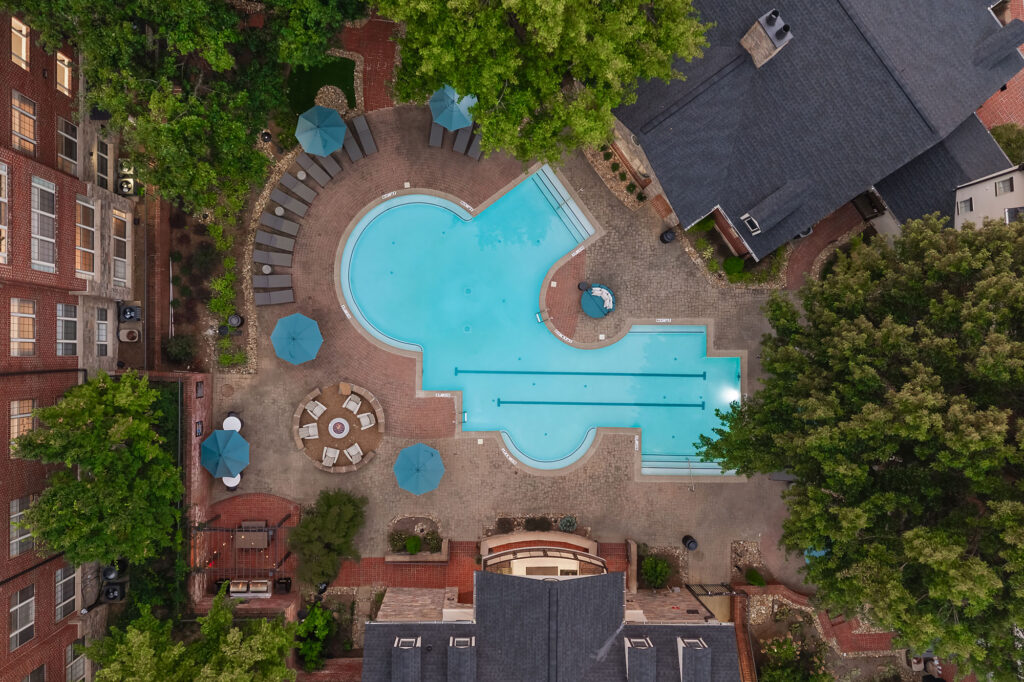 Aerial view of a courtyard with a curved swimming pool, patio furniture, and several blue umbrellas surrounded by buildings and trees.