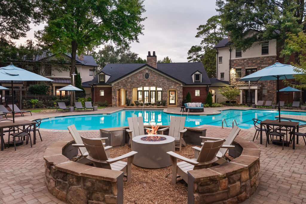 A circular fire pit surrounded by chairs on a stone patio overlooking a swimming pool and multi-story stone residential buildings.
