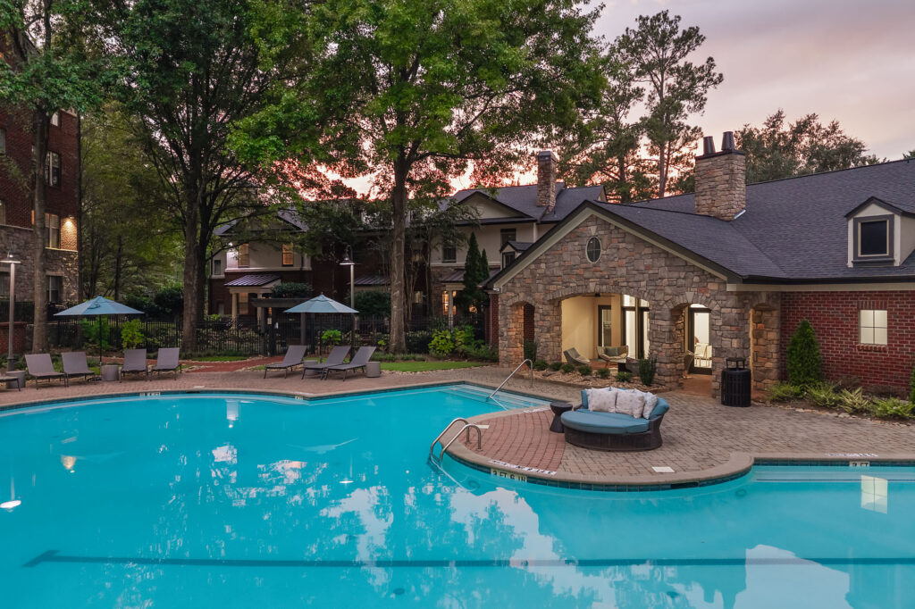 An outdoor swimming pool at dusk, featuring a stone clubhouse with a covered patio, lounge chairs, and a circular outdoor sofa on a brick pool deck.