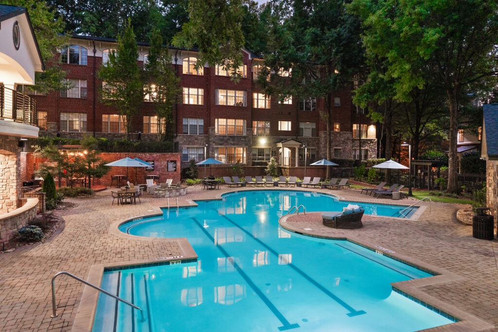 An outdoor swimming pool at night surrounded by a paved deck with lounge chairs, umbrellas, and a brick apartment building in the background.