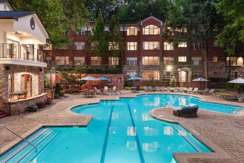 A large, multi-story brick apartment building overlooking an outdoor swimming pool with lounge chairs and patio tables.