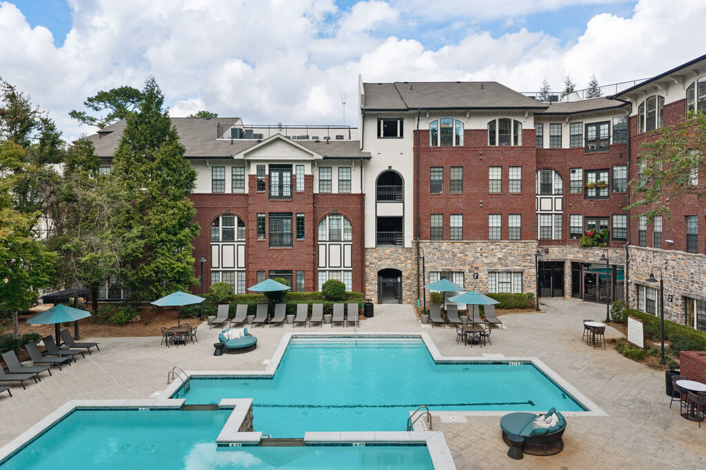A multi-level apartment complex with red brick and stone facades overlooking a swimming pool area equipped with lounge chairs, umbrellas, and small tables.