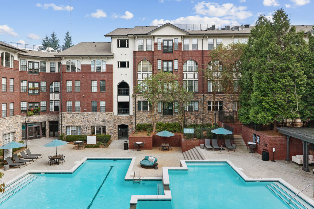 A multi-story brick apartment building overlooks an outdoor swimming pool with lounge chairs and patio tables.