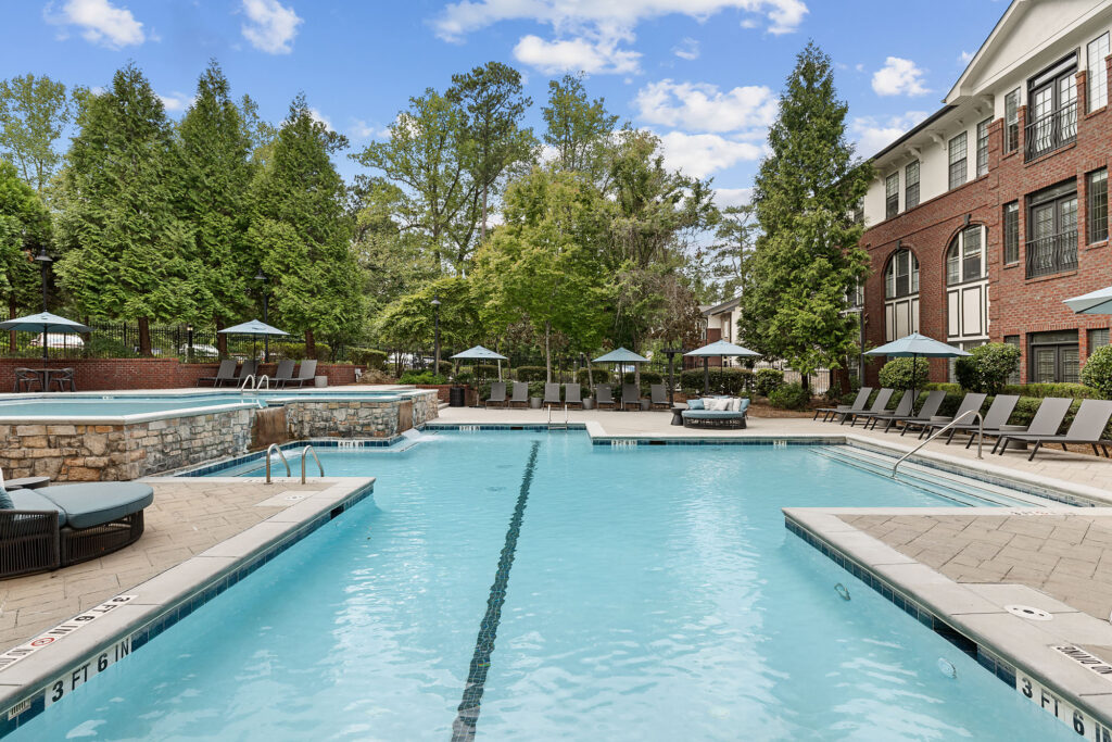 A large outdoor swimming pool at an apartment complex, surrounded by lounge chairs, umbrellas, and a brick building.