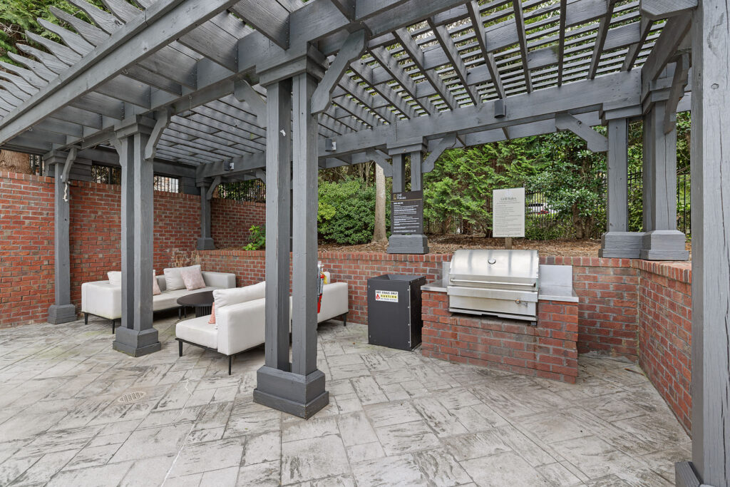 Outdoor patio area under a dark wooden pergola with a brick-surrounded grill, a trash bin, and two cream-colored sofas facing a small coffee table.