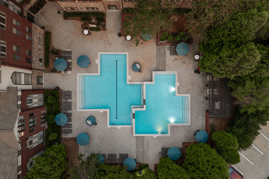 Aerial view of a cross-shaped swimming pool surrounded by a patio with lounge chairs, umbrellas, and surrounding brick buildings and trees.
