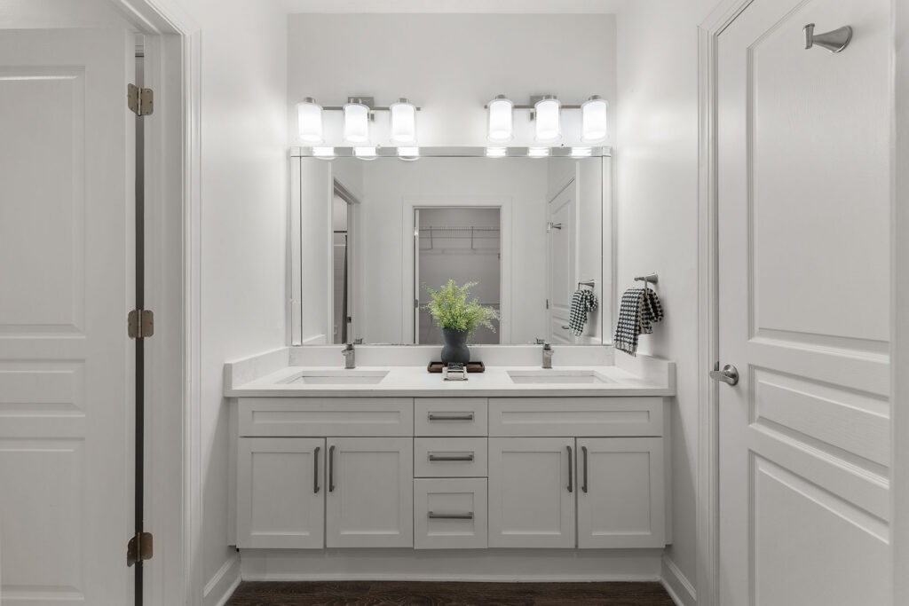 White double-sink bathroom vanity with silver faucets, a large mirror, and a central vase of greenery.