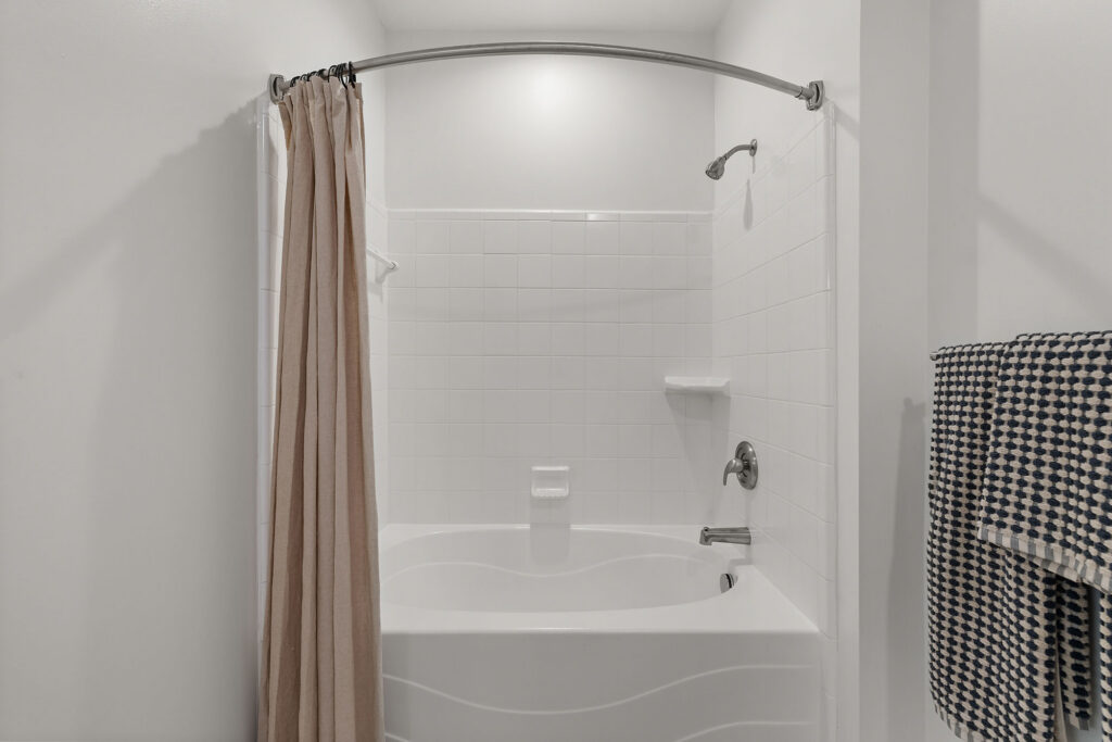 A white bathtub and shower combination with a beige curtain and patterned towels hanging on the wall.
