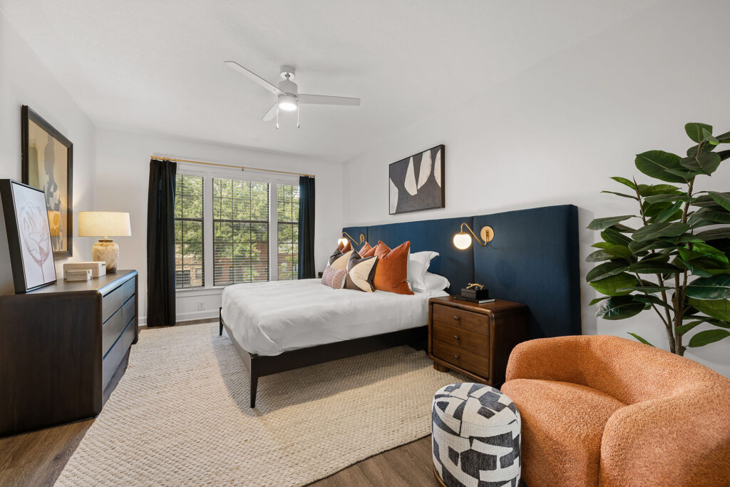 A bedroom featuring a queen bed with a tall navy headboard, two wooden nightstands, an orange accent chair with a patterned ottoman, and a large indoor plant.