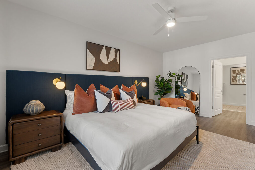 A bedroom with a king-sized bed featuring a navy blue headboard, orange and patterned pillows, two wooden nightstands, a ceiling fan, a potted plant, and an arched floor mirror.